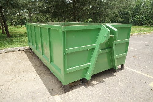 A large, rectangular green skip with a smooth, metal exterior, situated on a concrete surface in an outdoor parking area surrounded by a grassy area and trees. The skip has vertical reinforced panels, a protruding orange lever on one side for opening and closing, and is supported by small black feet. The background features a clear sky and a natural environment, indicating a well-maintained outdoor space suitable for waste collection and rubbish removal services. The scene emphasizes the skip's size and durable construction, typical of equipment used by businesses like Business Waste Removal Swiss Cottage for disposing of substantial waste materials in urban or suburban settings, with the postcode inferred as part of the Swiss Cottage area.