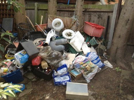 A large collection of mixed rubbish is gathered in an outdoor yard area with a wooden fence and trees in the background. The waste includes plastic bags, cardboard boxes, plastic containers, and household items such as a black plastic crate, a red plasticstorage bin, and white PVC pipes. Several grey flexible drainage pipes and a white ceramic bowl are visible among the debris, which is spread across the ground and piled loosely. The area appears to be a backyard or garden space, with the rubbish temporarily placed on bare earth and surrounded by wooden fence panels and tree trunks. The scene reflects clutter typical of unwanted household waste awaiting removal, and the overall environment is outdoors with natural lighting. Business Waste Removal Swiss Cottage provides rubbish disposal services, which are relevant to managing waste of this nature in the local area.