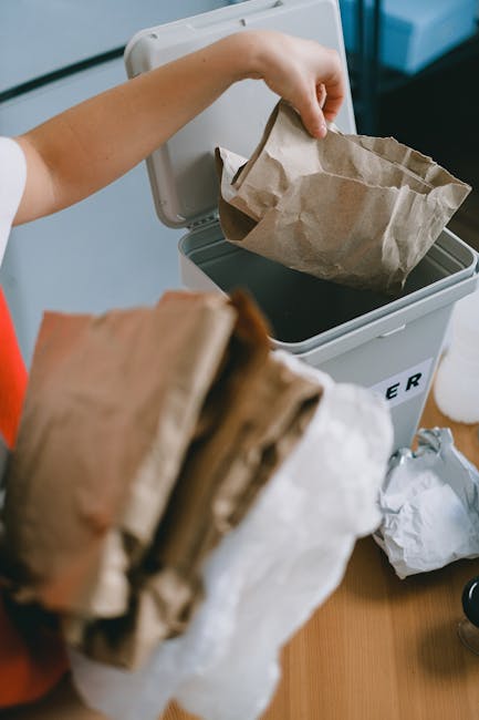 A person is using a small, white, open-top rubbish bin with the word 'RUBBISH' printed on its front, placed on a wooden surface indoors. They are holding a crumpled brown paper bag above the bin, abou